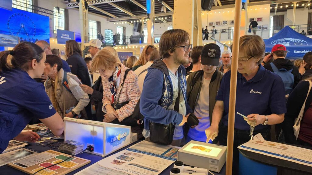 Preplnené podujatie v interiéri, kde sa ľudia zhromažďujú okolo stolov s vedeckými exponátmi. Účastníci sa zúčastňujú na praktických aktivitách, zatiaľ čo personál podáva výklad; viditeľné sú informačné materiály a osvetlené displeje.