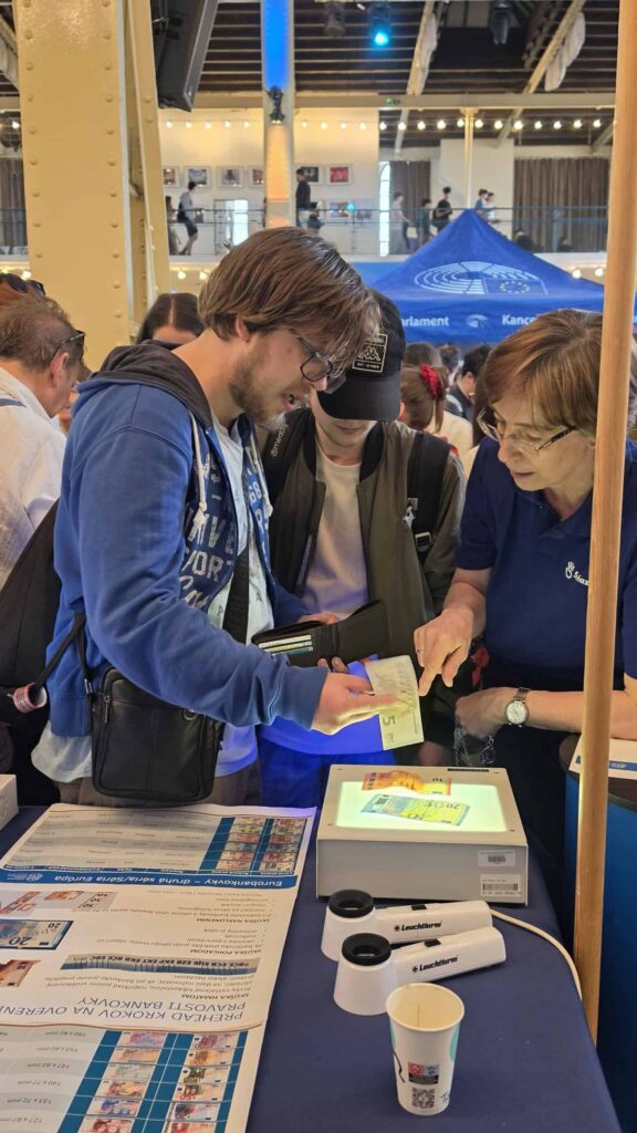 Dvaja mladí ľudia sa rozprávajú so ženou v informačnom stánku, ktorá skúma dokumenty pod osvetleným skenerom. V rušnej hale v pozadí je vidieť ďalších účastníkov a stánky.