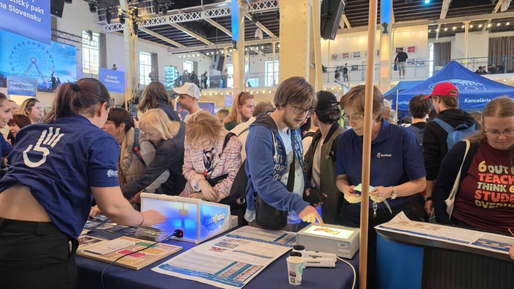 Preplnené podujatie v interiéri s ľuďmi komunikujúcimi pri informačných stánkoch. Účastníci skúmajú materiály a muž používa dotykový displej. Zamestnanci v modrých košeliach pomáhajú návštevníkom. Transparenty a svetlá zdobia živé, priestranné miesto.