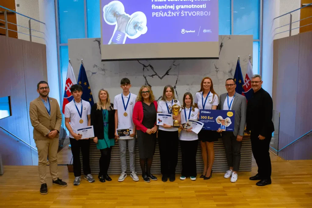 Skupina ľudí, medzi ktorými sú aj študenti s medailami, stojí v rade s oceneniami a veľkým šekom na 300 eur a pózuje na fotografiu v interiéri so slovenskými vlajkami a transparentom s cenami za nimi.