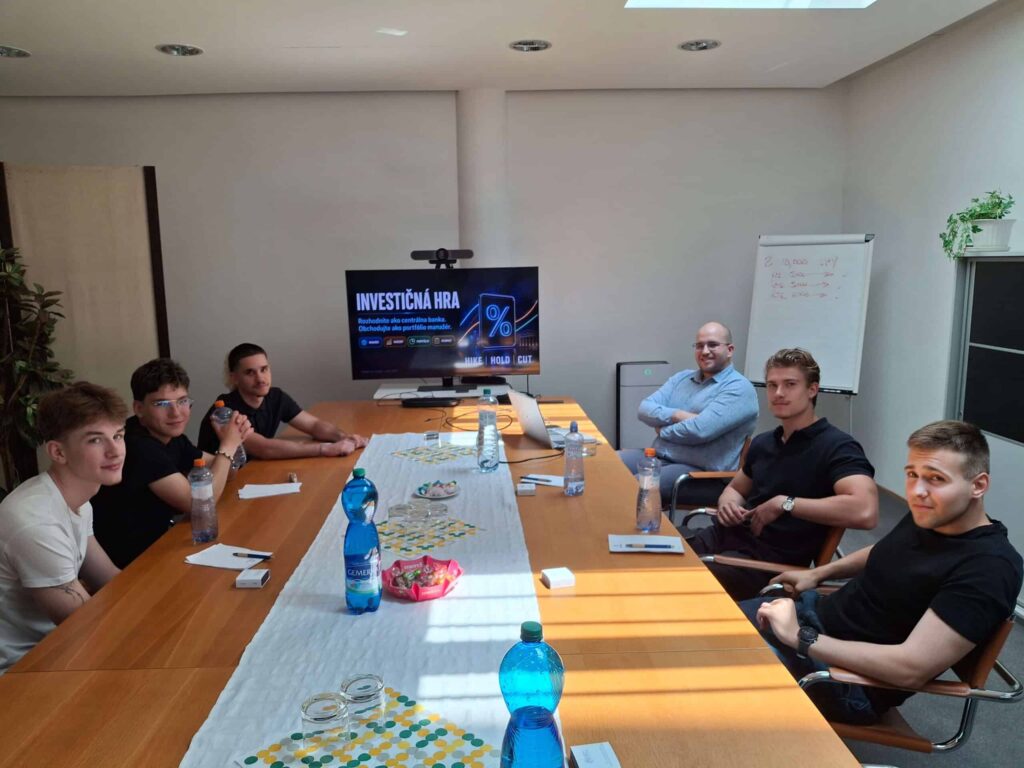 Šesť mladých mužov sedí okolo konferenčného stola s fľašami s vodou, blokmi na poznámky a občerstvením. Na obrazovke sa zobrazuje hra „Investičná hra“ a finančné grafy. V pozadí je biela tabuľa a veľké okná.