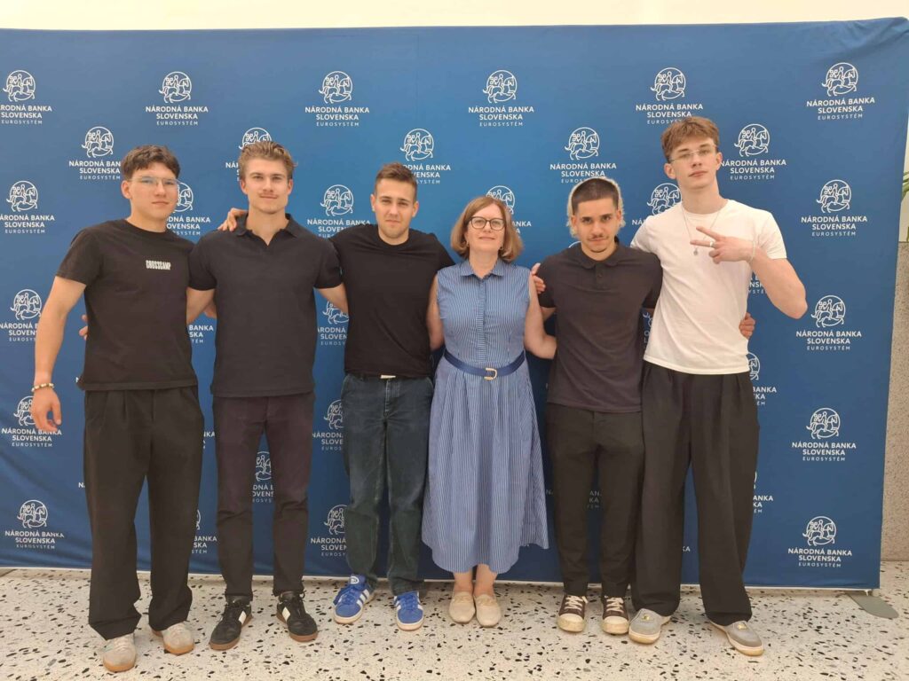 Šesť ľudí stojí v rade a usmieva sa do fotoaparátu pred modrým pozadím s bielymi logami a nápisom „Národná banka Slovenska“. V skupine je jedna žena a päť mužov.