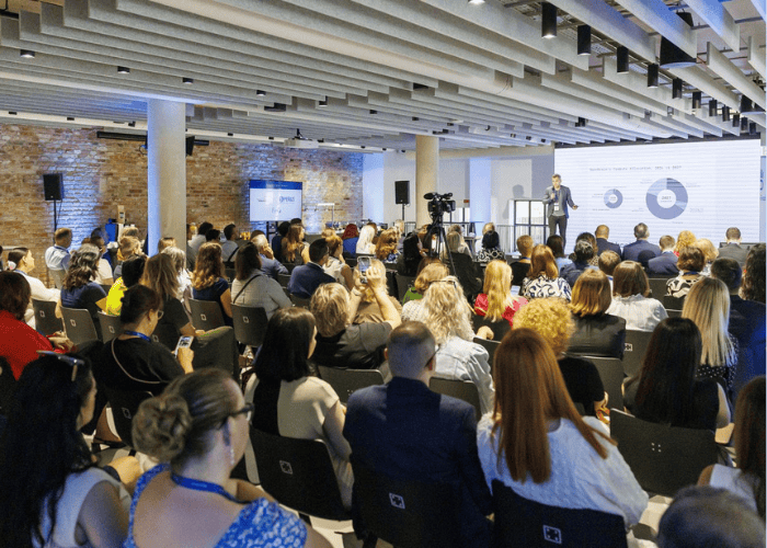 Konferencia Príklad pre vzdelávanie - V modernej konferenčnej miestnosti s odhalenými tehlovými stenami a stropnými trámami sedí početné publikum v radoch oproti rečníkovi, ktorý na obrazovke prezentuje vizualizácie údajov.