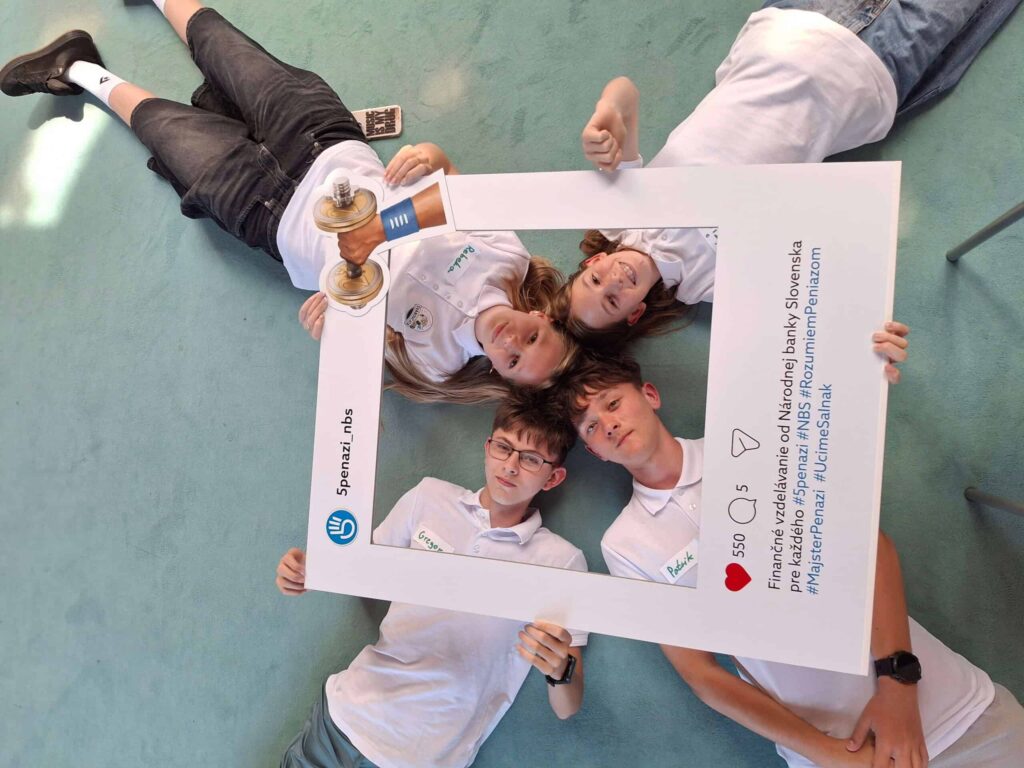 Štyria mladí ľudia v bielych košeliach ležia na zelenej podlahe, hlavy pri sebe, držia veľký rám v štýle Instagramu s malou trofejou a hľadia do kamery.