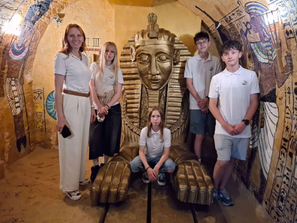 Päť ľudí v bielych košeliach pózuje okolo veľkej zlatej sochy faraóna v miestnosti s egyptskou tematikou, ktorej steny zdobia hieroglyfy; jedna osoba sedí pred sochou so skríženými nohami, zatiaľ čo ostatní stoja.