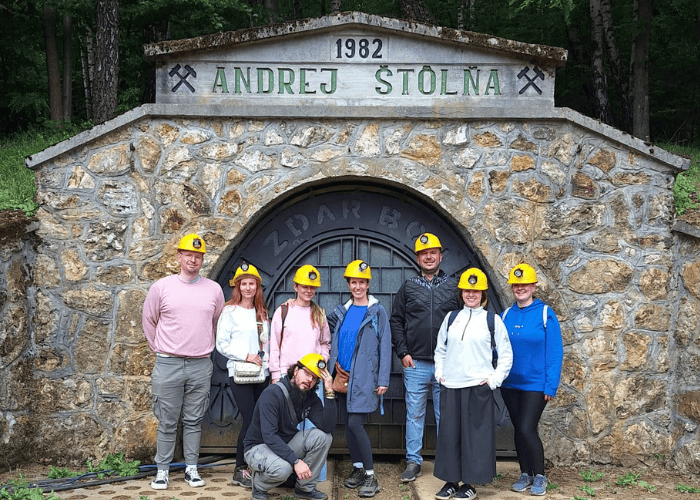 Osem ľudí v žltých ochranných prilbách pózuje pred vchodom do kameňolomu s nápisom „Andrej Štolna 1982“, obklopeného zeleňou. Niektorí stoja, jeden z nich kľačí vpredu, všetci sa usmievajú do fotoaparátu.
