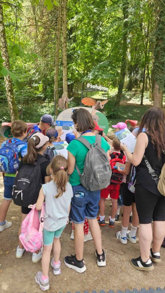 Skupina detí a dospelých s batohmi sa zhromažďuje okolo informačnej tabule v zoologickej záhrade, obklopenej stromami a zeleňou. Na tabuli je nápis #setulzoo a sú na nej vyobrazené zvieratá.