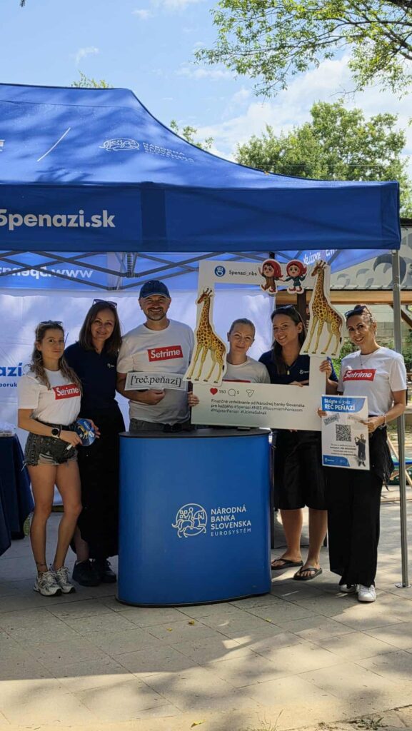 Pred modrým stánkom s nápisom v slovenčine stojí šesť ľudí, ktorí držia vystrihnuté postavy žiráf a propagačné tabule. Stánok je umiestnený vonku pod stromami a sú na ňom viditeľné tričká a propagačné materiály so značkou.