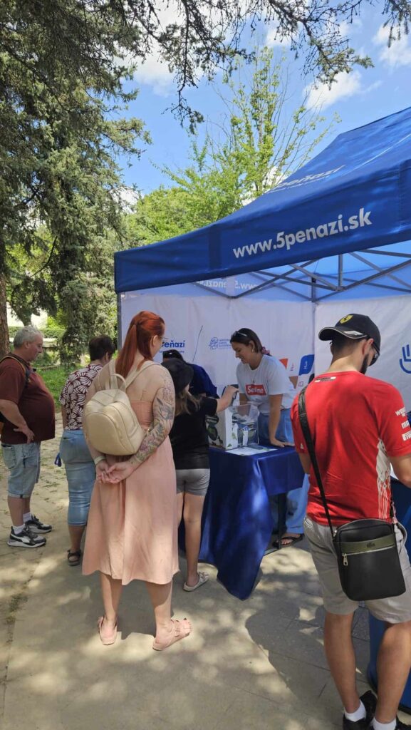 Ľudia stoja v rade pri stánku pod modrým stanom s nápisom 5penazi.sk. Zamestnanec za stolom sa rozpráva s návštevníkmi. V pozadí sú vidieť stromy a slnečná obloha.