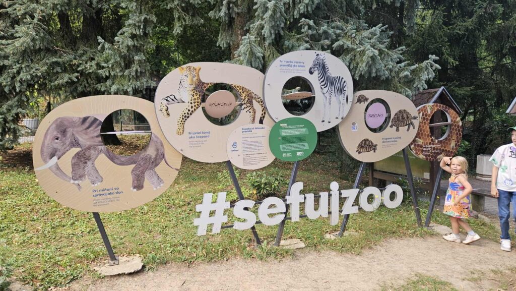 Deti pózujú pred fototabulou zo zoologickej záhrady s ilustráciami zebry, tigra, slona a žirafy, na ktorej je veľkými písmenami napísané #sefuiZoo. V pozadí sú stromy a tráva.