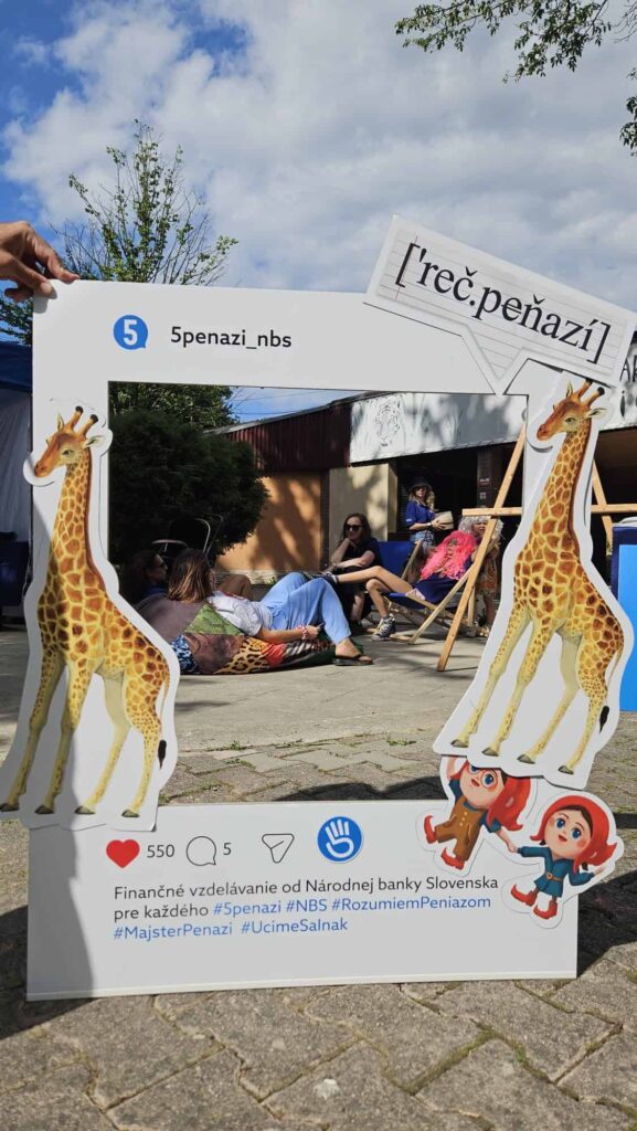 Ľudia sedia na ležadlách vonku za dekoráciou v podobe fotorámika, ktorý je vyzdobený kreslenými žirafami a deťmi. Slovenský text na rámiku propaguje finančné vzdelávanie, ktoré organizuje Národná banka Slovenska.