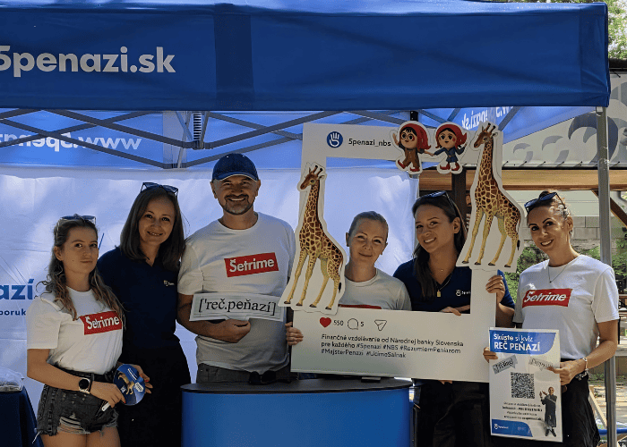 Šesť ľudí stojí pod modrým stanom s nápisom v slovenčine, usmievajú sa a držia v rukách vystrihnuté postavy žiráf, reklamné tabule a QR kódy, čím propagujú finančné vzdelávanie. Majú na sebe ležérne tričká a zdá sa, že sa nachádzajú na podujatí pod holým nebom.