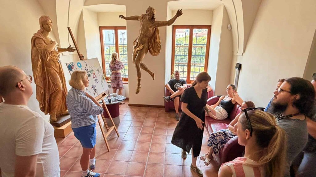 Ľudia sa zhromažďujú v slnkom osvetlenej miestnosti so sochami; niektorí sedia, iní stoja a rozprávajú sa. V miestnosti sú veľké okná, maliarsky stojan s plagátom a podlaha pokrytá terakotovými dlaždicami.